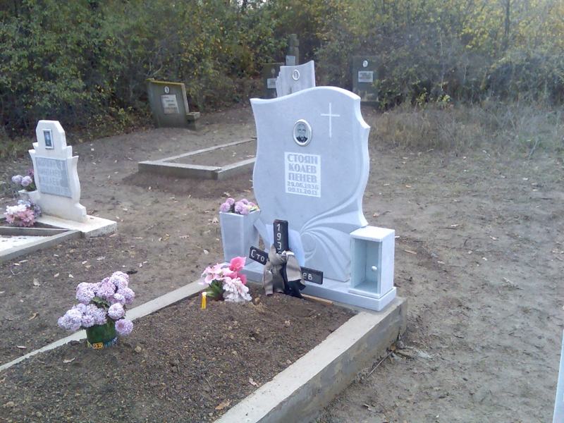 Паметник от светлосив мрамор модел колелото на живота, с вдълбан кръст, черно-бяла елипсовидна порцеланова снимка, надпис от релефни букви, постамент, ваза и фенер от плочки, бетонен бордюр.